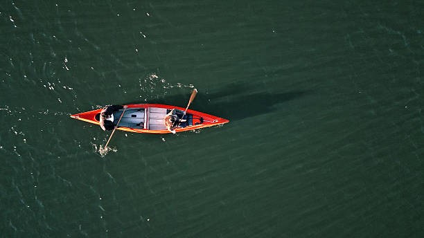 Aerial shot of a couple paddling an orange canoe on calm waters, enjoying a serene weekend activity. The scene highlights their active lifestyle, connection with nature, and the joy of outdoor adventures and shared moments.