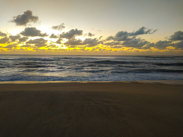 Dramatic golden light breaks through heavy clouds over rhythmic ocean waves and dark sand. This serene coastal landscape evokes hope, perfect for travel, wellness, and nature themes.