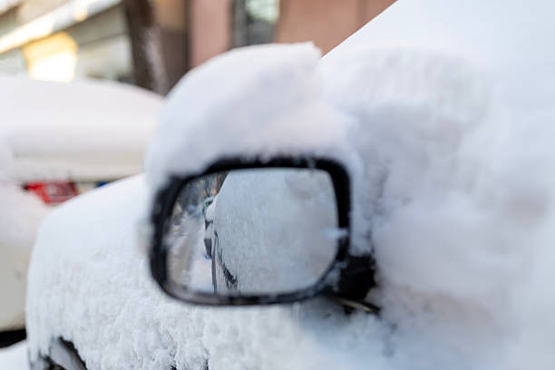 Automobile side mirror and car body completely covered in a thick blanket of fresh white snow, reflecting a wintery, frozen street scene during a cold temperature day