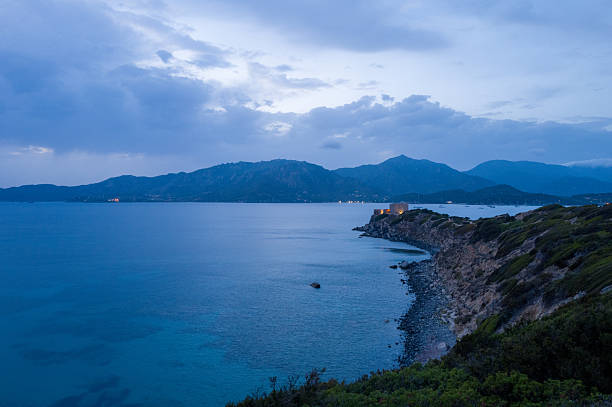 Serene twilight view of Capo Carbonara's rugged shoreline with a solitary fortress illuminated above calm blue waters and distant mountain silhouettes under a dramatic cloudy sky.