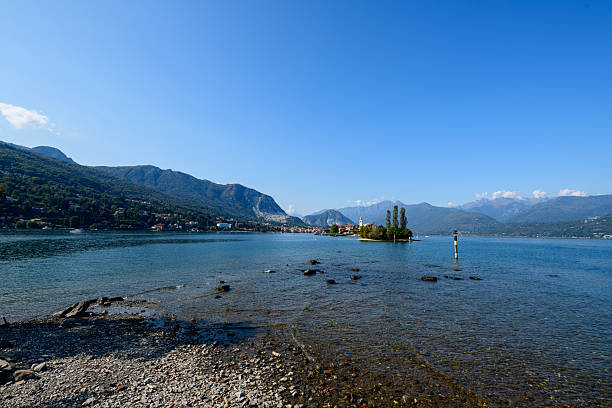 Clear blue water and a rocky shoreline frame a distant view of Isola Bella, set against the forested hills and mountains of Lake Maggiore in northern Italy. The tranquil scene features open sky and gentle ripples along the lakeshore.