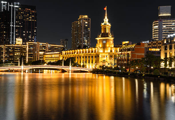 Illuminated night view of the historic Shanghai Post Office along Suzhou Creek in Shanghai, China. Warm golden architectural lighting reflects across the calm river, blending early 20th-century neoclassical design with the modern city skyline and bridges, capturing Shanghai’s layered urban history after dark.