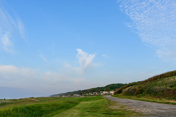 Winding dirt road leads through green grass and gentle hills beneath a wide sky with wispy cirrus and altocumulus clouds in Normandy. Soft evening light creates a calm, open atmosphere.