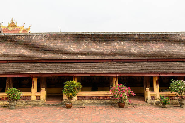 Long wooden gallery with sloped tiled roof and yellow columns lines the brick courtyard at Wat Si Saket temple in Vientiane, Laos. Potted flowering plants and subtle decorative details create a tranquil, historic atmosphere under soft daylight.