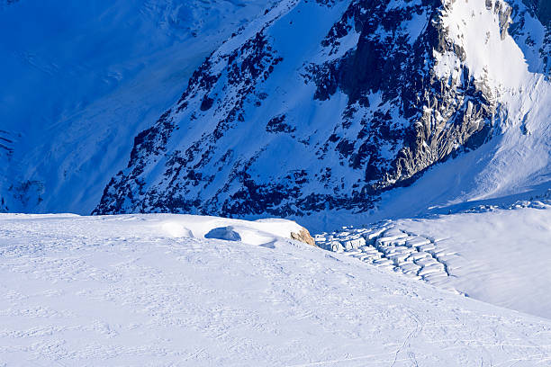 Deep blue shadows stretch across a pristine snow slope above a fractured glacier in the Mont Blanc massif, with rugged mountain textures in the background.
