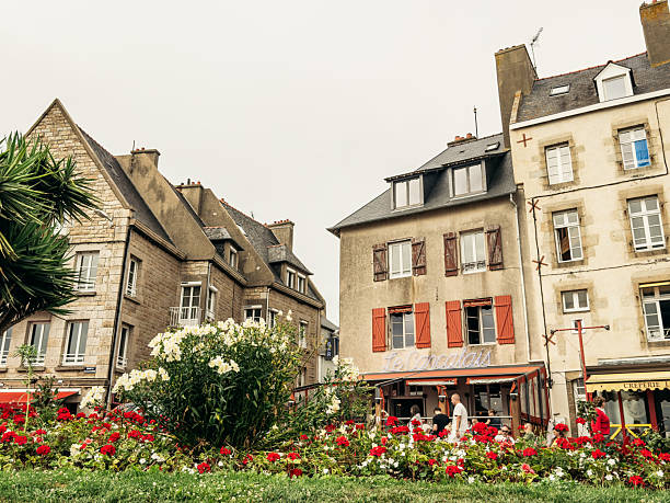 People gather near cafes and flower beds among historic stone buildings in Saint-Malo, Brittany, capturing everyday summer life in a coastal French town.