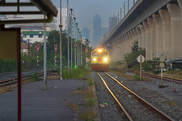 Bangkok, Thailand - January 11, 2026: a diesel railcar train with bright headlights is seen approaching the platform at Hua Mak railway station during the early morning hours. the scene captures the atmospheric contrast between traditional rail transport and the modern elevated railway structure of the airport rail link in the background.