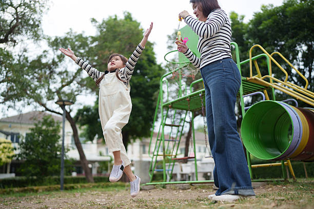 Mother creating bubbles while her daughter jumps and plays outside. Lifestyle scene focused on energy and childhood happiness.