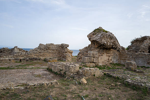 Ancient stone ruins with rough, eroded textures stand on open ground at the Tharros archaeological site near Cabras, Sardinia. Subtle daylight highlights the layered masonry and hints of the sea in the distance.