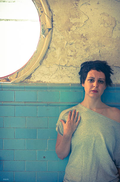 Model posing for a portrait in an abandoned house expressing emotions: sadness, loneliness, self-absorption, simplicity, nostalgia, thoughtfulness, grief,in front of an old wall with peeling paint and next to a broken window