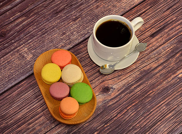 Cup of black coffee on a saucer with a spoon and a wooden plate with six multi-colored macaroons on a wooden table.