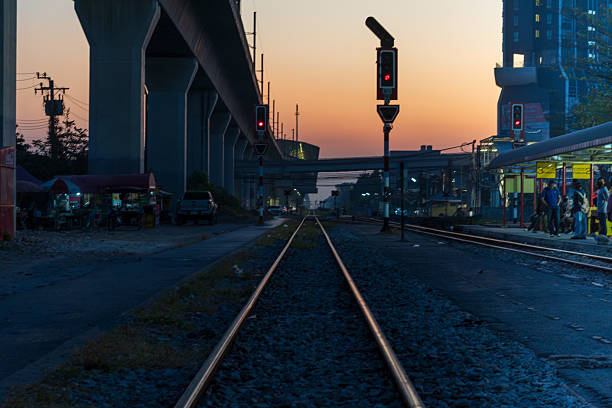 Bangkok, Thailand - January 11, 2026: a group of commuters is seen waiting for a train on the platform at Hua Mak railway station at 6:40 am. the scene captures the silhouette of passengers against a vibrant sunrise sky, with the modern airport rail link structure visible in the background.