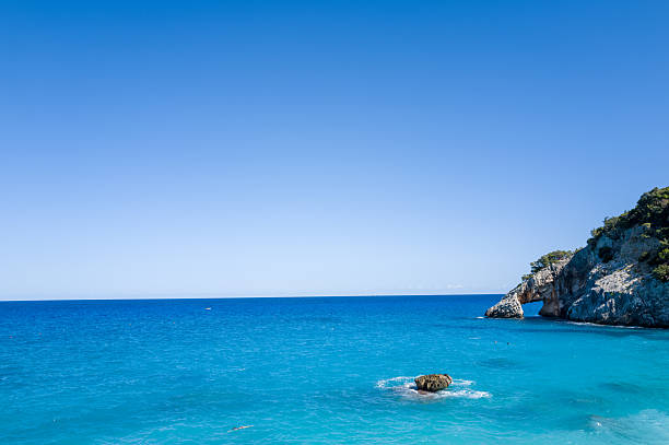 A striking rocky promontory juts into vibrant blue Mediterranean waters at Cala Goloritze, Baunei, Sardinia. The clear sky and sunlight highlight the crisp contrast between the rugged coastline and the tranquil sea.