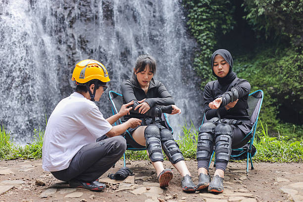 Group Of Friends Gearing Up For Canyoning Trip In Front Of Waterfall With Muslim Woman Helps Adjust The Leg Straps Of Her Friend's Harness
