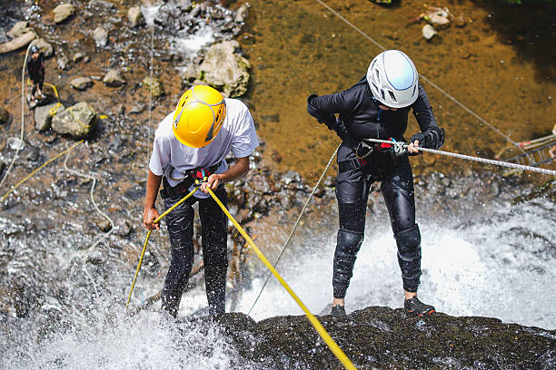 High Angle Shot Of Muslim Woman In Hijab And White Helmet Cautiously Descending Waterfall Alongside Her Male Companion In Yellow Helmet