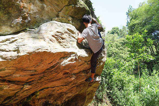Low Angle Shot From Behind Of Male Rock Climber Reaching For The Top Edge Of Large Boulder In Lush Forest
