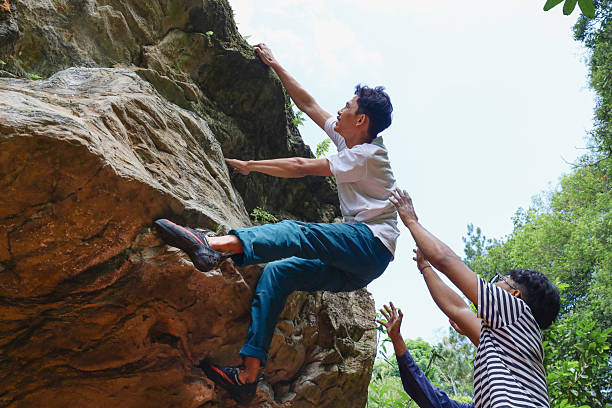 Low Angle Shot Of Male Rock Climber Negotiating Difficult Overhang While Dedicated Spotter Stands Below With Arms Raised To Ensure Safety
