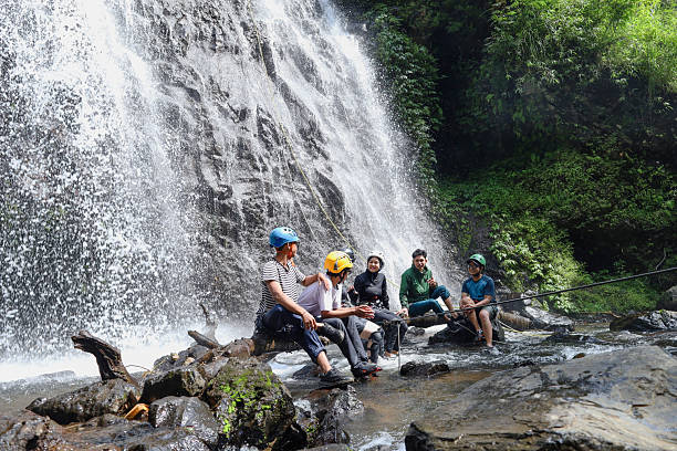Group Of Friends And Guides Resting On Log At The Base Of Massive Waterfall