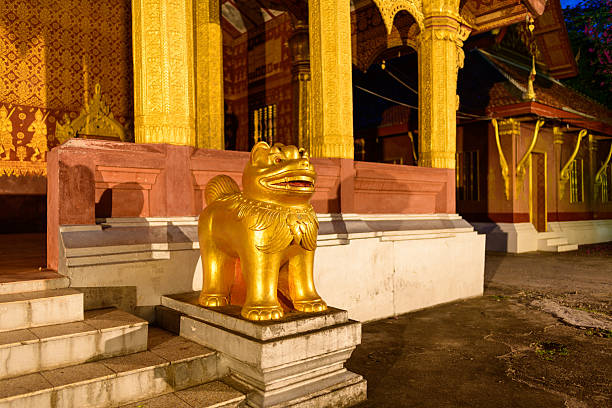 A golden guardian lion statue stands at the entrance steps of an ornate Buddhist temple in Luang Prabang, Laos. The richly decorated columns and intricate patterns are illuminated by warm evening light, highlighting Southeast Asian architectural heritage.