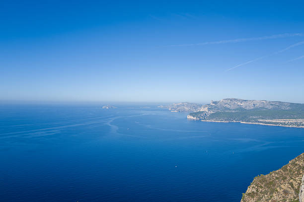 Expansive view of the deep blue Mediterranean Sea with rugged limestone cliffs and green hills stretching along the Cassis coastline under a clear sky. The tranquil scene is bathed in bright daylight and offers a sense of openness and natural beauty.