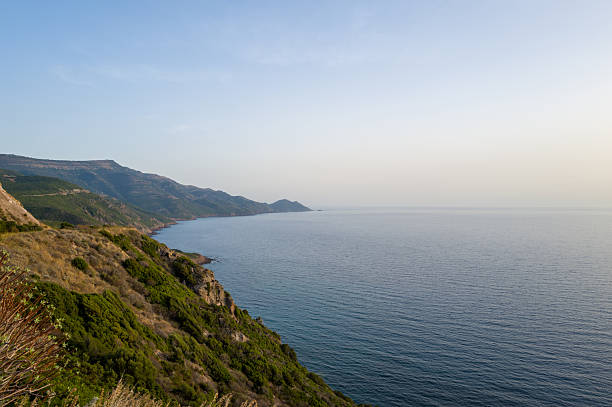 A tranquil view of the coastline between Alghero and Bosa, Sardinia, with green hillsides meeting the calm blue Mediterranean Sea under soft, clear evening light.