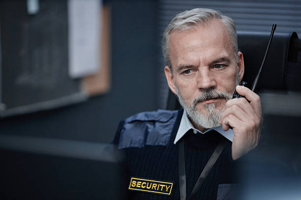 Middle aged Caucasian man with gray beard wearing security uniform sitting at desk, holding walkie talkie, looking thoughtfully to side, working in control room monitoring situation