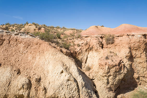 Sunlit ochre and beige rock formations display deep cracks and rugged textures in the arid landscape of Bardenas Reales, Spain. Sparse grasses and a vivid blue sky add contrast to the dry, eroded terrain.
