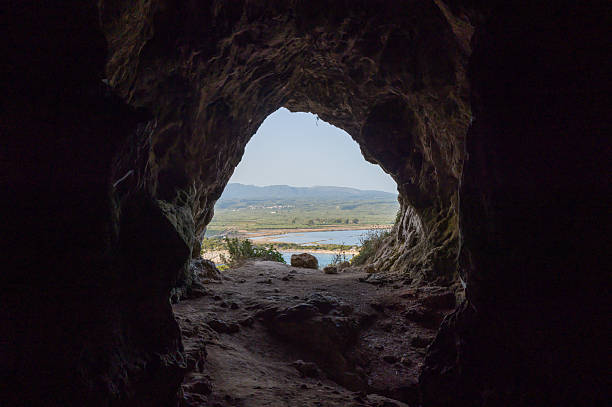 View from inside a rocky cave looking out toward a sunlit lagoon, green fields, and distant hills in Messenia, Peloponnese. Natural textures and earthy tones frame the bright landscape beyond.