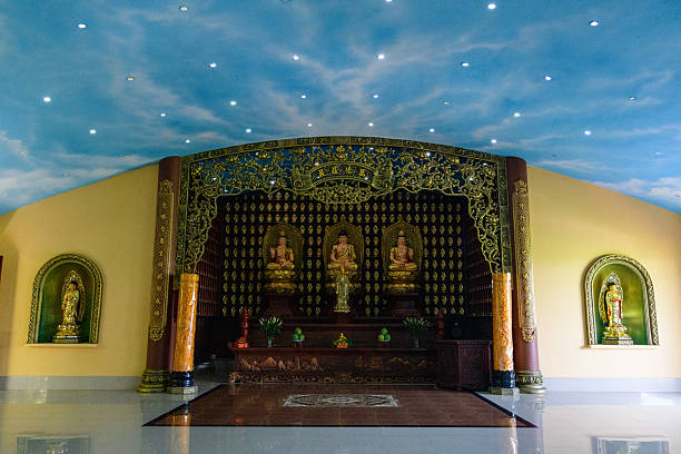 Three golden Buddha statues sit on a dark wooden altar framed by intricate gold arch detailing, with a painted sky ceiling and glossy floors creating an uplifting, tranquil temple ambiance.