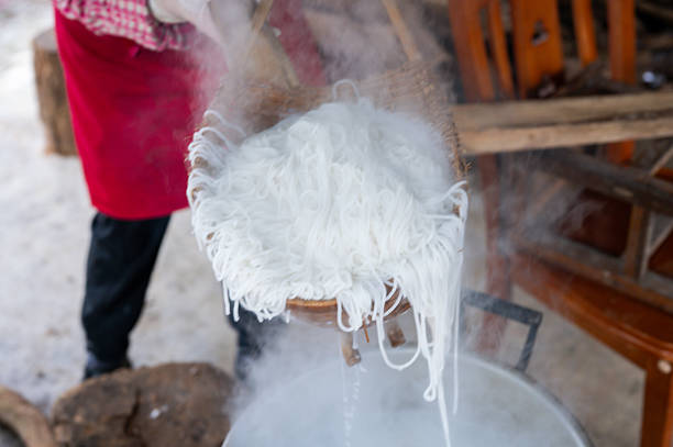 A chef skillfully lifts freshly cooked rice noodles from a boiling pot using a large bamboo basket, highlighting the intricate process of preparing traditional Asian cuisine in a bustling kitchen.
