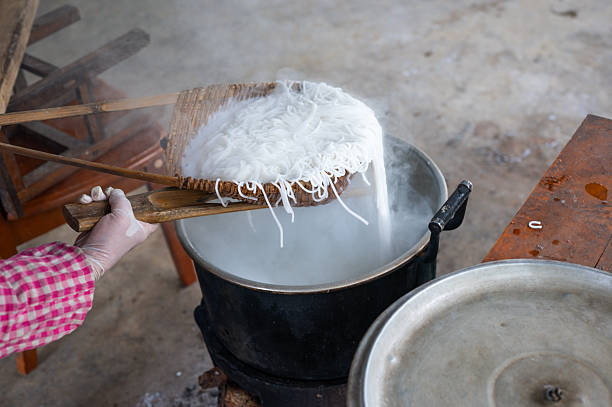 A chef skillfully lifts freshly cooked rice noodles from a boiling pot using a large bamboo basket, highlighting the intricate process of preparing traditional Asian cuisine in a bustling kitchen.