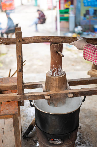 An adult worker presses rice dough through a manual lever as fresh noodles fall directly into boiling water above an open fire. The scene captures a traditional cooking method used in old-fashioned noodle preparation.