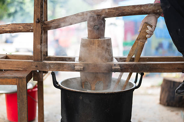 An adult worker presses rice dough through a manual lever as fresh noodles fall directly into boiling water above an open fire. The scene captures a traditional cooking method used in old-fashioned noodle preparation.