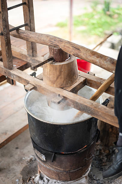 An adult worker sits on a manual lever press to apply pressure as rice dough forms noodles that drop into boiling water above an open flame. The scene illustrates a traditional and labor-intensive noodle cooking process.