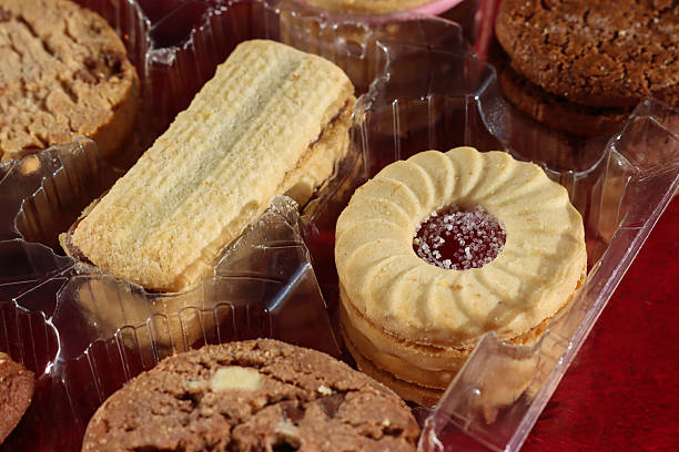 Stock photo showing close-up, elevated view of an assortment of biscuits in a disposable, single use plastic tray inset of a biscuit tin, pictured against a mottled red background.