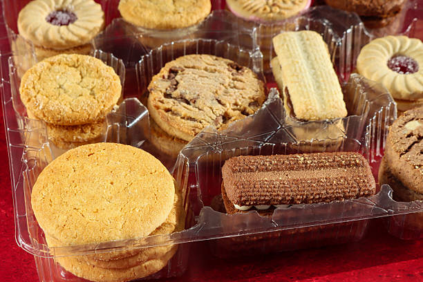 Stock photo showing close-up, elevated view of an assortment of biscuits in a disposable, single use plastic tray inset of a biscuit tin, pictured against a mottled red background.