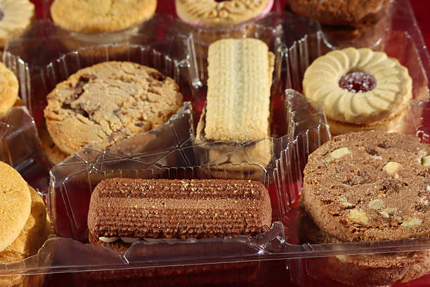 Stock photo showing close-up, elevated view of an assortment of biscuits in a disposable, single use plastic tray inset of a biscuit tin, pictured against a mottled red background.