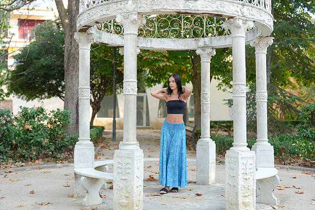 A woman is standing in front of a stone pavilion. She is wearing a black tank top and blue pants