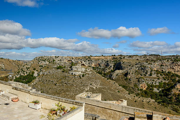 A spacious stone terrace with potted plants overlooks the rugged canyon landscape of Matera, Italy, under a bright blue sky dotted with fluffy clouds. The sunlit scene highlights the textures of ancient masonry and rocky hills.