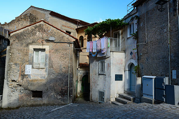 Colorful laundry hangs on a line between weathered stone buildings in a quiet Maratea street. Textured facades, cobblestone pavement, and soft daylight create a lived-in Mediterranean atmosphere.