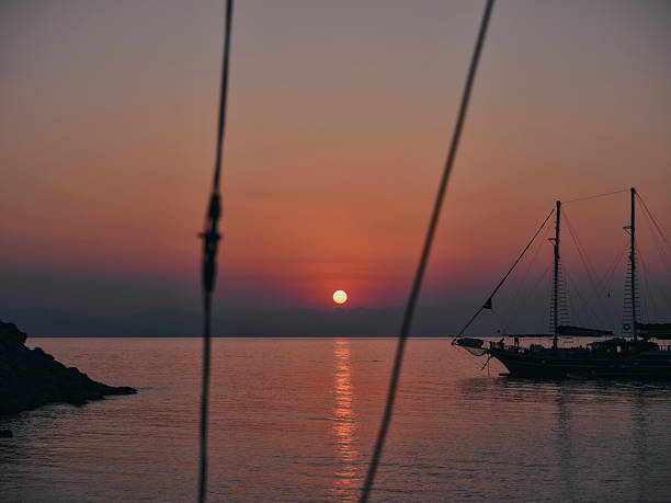 Golden sunset over the sea with a traditional wooden sailboat silhouette framed by ship ropes.