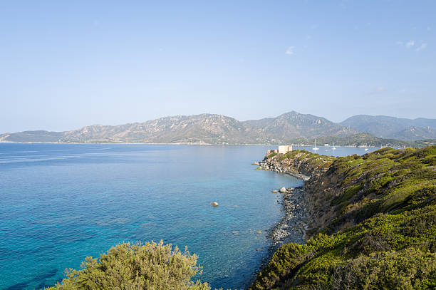 Bright daylight reveals the clear turquoise waters and rugged headland of Capo Carbonara, with a solitary fortress overlooking the Mediterranean and distant mountain ranges under a clear blue sky.