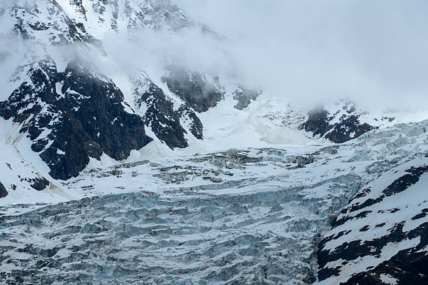 Layered blue ice of Glacier des Bossons sprawls beneath steep, snow-draped cliffs, with misty clouds partially obscuring the dramatic alpine landscape in the French Alps.