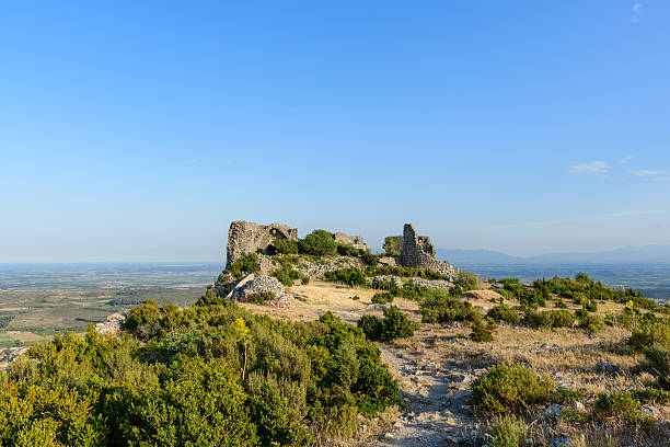Crumbling stone walls of an ancient fortress crown a rugged hilltop, surrounded by dry grass and low shrubs, with expansive valley views under a clear blue sky near Opoul Perillos.