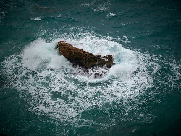 Aerial-like view of a dark rock in the sea being hit by white foamy waves.