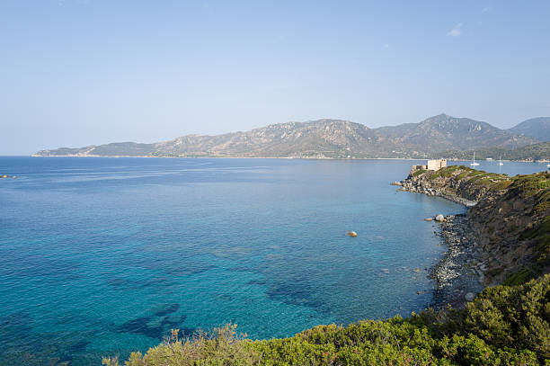 Clear turquoise waters stretch along the rocky coastline of Cala Caterina, with an old stone watchtower perched on the headland and rugged mountains rising in the background under a bright sky.
