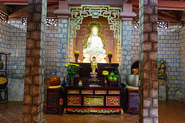 Large white Buddha sculpture sits above a richly decorated altar with offerings, flowers, and intricate woodwork, framed by stone pillars and patterned brick walls in a tranquil temple interior.