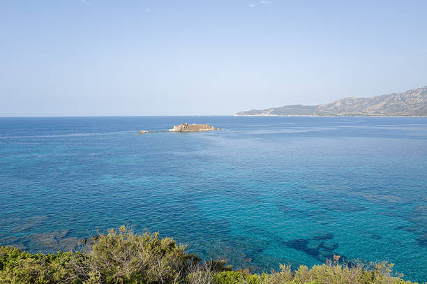 Vivid turquoise waters surround a small rocky islet off the coast of Capo Carbonara, with distant mountains and a bright sky creating a peaceful Mediterranean scene.