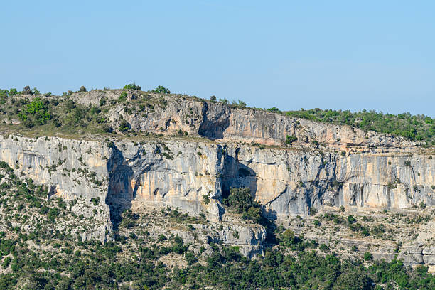 Broad limestone cliffs with rugged textures and scattered greenery rise steeply beneath a clear blue sky at Cirque de Navacelles. The scene is bright, natural, and dramatic.