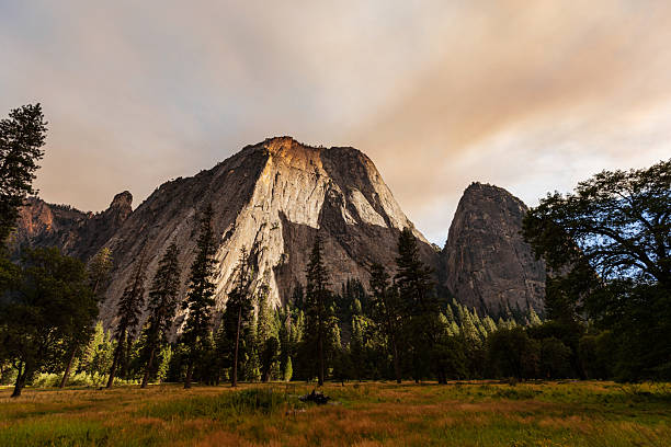 Granite cliffs and forest meadow in Yosemite National Park illuminated by warm wildfire haze, creating dramatic light and smoky atmosphere over the iconic mountain landscape.